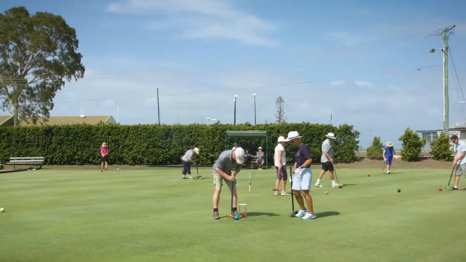 Players walking out onto a wide, sunny croquet lawn lined by a hedge