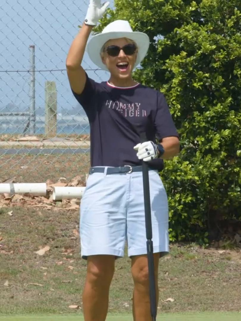 A smiling player in a sun hat waving from the edge of a croquet lawn